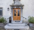The image shows the entrance to a building with a decorative stone facade. The entrance has a set of double wooden doors with intricate metal designs. There is a stone staircase leading up to the entrance, and potted plants on either side. A lantern is mo