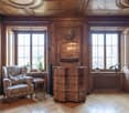Ornate wooden paneled walls and ceiling, large windows, antique wooden dresser, armchair, potted plants