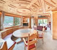 Spacious living room with large windows offering a scenic mountain view, ornate wooden ceiling with decorative moldings, round wooden dining table, red leather chairs, and a cozy seating area with sofas.