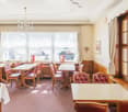 This image shows a traditional, cozy dining room with wooden furniture and decor. The room has large windows that let in natural light, a crystal chandelier, and red tufted leather chairs around wooden tables. The overall atmosphere appears to be that of