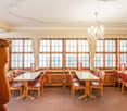Large dining room with wooden furniture, including a long table with red upholstered chairs, a chandelier, and large windows providing natural light.