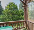 Balcony with wooden railing, green round table, potted plants, view of trees, and colorful glass window