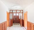 The image shows a spacious entrance hall with a high ceiling and ornate plaster moldings. The walls are lined with wood paneling, and the floor is covered in a patterned tile. The main entrance features a set of large, wooden double doors with stained gla