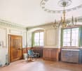 Large room with wooden paneling, ornate fireplace, chandelier, wooden floor, radiator, and a wooden chair.