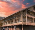 A multi-story wooden building with a large balcony or terrace running along the front, set against a dramatic sunset sky with orange and pink clouds.