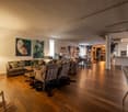 Open living space with wooden floors, large couch, coffee table, dining table, chairs, lamps, paintings, kitchen area