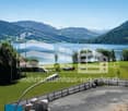 House with a view of a lake and mountains, surrounded by a green field, with a barn and a fence