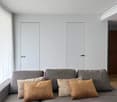 Gray couch with four pillows, white curtain on the left, white wall, two white doors, brown door to the right