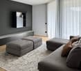 modern living room, gray walls, large gray sofa, two gray ottomans, gray rug, gray wall cabinets, television mounted on the wall, white curtains