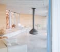 Minimalist interior with large white couches, large glass windows, white marble flooring, hanging cylindrical fireplace, white walls, white chairs, white dining table, dried plants