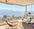 Spacious living room with floor-to-ceiling glass windows offering a panoramic view of the surrounding mountains and landscape. The room features a comfortable sofa, armchair, and a wooden coffee table. The balcony provides an outdoor seating area with a s