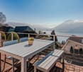 Spacious outdoor terrace with wooden deck, dining table and benches, overlooking a scenic lake and mountain landscape. The terrace is surrounded by lush greenery and has a panoramic view of the natural surroundings.
