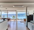 Spacious open-plan living area with large windows offering a panoramic view of a lake and surrounding hills. The room features a modern kitchen with white cabinets, a dining table with chairs, and a comfortable sofa. The decor includes potted plants, deco