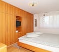 A bedroom with wooden floor, a double bed, wooden wardrobe, shelves, a window with curtains, and decorative heart-shaped wall decor.