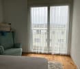 Bedroom with large windows, white curtains, green armchair, wooden floor, and a decorative pillow on the bed