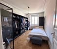 A single bed with storage below, wardrobe, parquet flooring, white walls, large window, white sheets and pillows