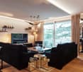 Living room with black sofas, glass coffee table, dining table, television, modern lighting fixtures, balcony visible through the sliding doors, large windows, wooden flooring, beige walls