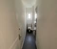 Hallway leading to bathroom, white walls, grey floor, white toilet, white sink, small window, recessed lighting