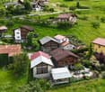 Aerial view of a picturesque village nestled in a lush, green valley. The village consists of traditional Alpine-style houses with red tile roofs, surrounded by well-maintained gardens and orchards. There are several small outbuildings and a few vehicles