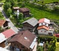 Aerial view of a picturesque village nestled in a lush, green landscape. The village features a mix of traditional wooden and stone buildings with red tile roofs, surrounded by well-maintained gardens and greenery. A winding road runs through the village,