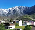 The image shows a picturesque mountain village nestled in a valley, with snow-capped peaks in the background. The buildings have a traditional alpine architecture style, with colorful facades and roofs. The landscape is lush and green, with trees and vege