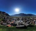 This image shows a panoramic view of a mountainous town or city. The town is surrounded by mountains and has a mix of residential and commercial buildings with red roofs. The sky is clear and blue, with the sun shining brightly overhead.