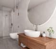 modern bathroom with a glass shower enclosure, toilet, large round mirror, wooden vanity, white porcelain sink, shower head, towels, lotion bottles, flowers