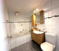 white tiled walls and floor, bathtub with shower, sink, toilet, wooden cabinet with shelves, mirror with lights