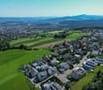 Aerial view of a suburban neighborhood with houses and a city in the distance, surrounded by green fields and trees, with a clear blue sky.