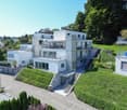 Modern architectural design, white exterior, multiple balconies, green hillside, glass windows, landscaped entrance with stairs