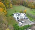 Aerial view of a large white house with two garages, surrounded by a grassy field and trees.