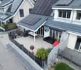 Aerial view of houses with solar panels on the roofs, modern architecture, fenced garden, outdoor patio with table, chairs, and a barbecue, solar-powered water heater, a concrete walkway.