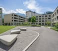 Multi-story apartment buildings with balconies, surrounded by a landscaped courtyard with gravel paths and benches. The buildings have a modern, minimalist design with large windows.