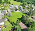 Large open green plot of land, surrounded by modern houses and greenery, with a road and pathway visible in the middle.