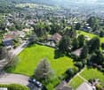 Aerial view of a large green plot of land with trees and shrubs, a neighborhood in the background, and a view of the mountains.