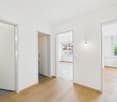 Empty room, wooden floor, white walls, open doors, visible toilet, white tiles, radiator, windows, hallway