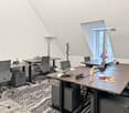 The image shows a modern office space with several desks, chairs, and office equipment. The desks have a wooden finish and there are adjustable lamps and a computer monitor on each desk. The floor is covered with a patterned carpet. There is also a small