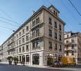 Multi-story building with ornate architectural details, balconies, and shops on the ground floor. The building has a classic European style with arched windows and decorative elements. There is a delivery van parked on the street in front of the building.