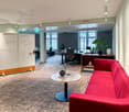 Modern office with white walls and carpeted floor, large windows, white round table, red sofa, multiple desks, plants, lamps, whiteboards