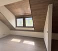 Spacious attic room with slanted wooden ceiling, large windows overlooking the outdoors, and hardwood flooring. The room has a modern, minimalist design with clean lines and neutral colors.