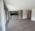 Empty room with wooden floor, white walls, kitchen with cabinets, microwave, oven, and a sliding glass door.