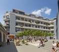 Multi-story apartment building with balconies, surrounded by trees and a public plaza with people sitting and socializing. The building has a modern, gray and white facade with large windows.
