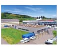 Large commercial building with a green roof, surrounded by a parking lot with several vehicles. The building appears to be a warehouse or distribution center, with loading docks and trucks visible. The surrounding landscape includes forested hills in the