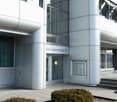 modern office building with sleek silver and glass exterior, Steinackerstrasse 29 address sign, double doors, windows with retractable awnings, circular security light above the entrance, and a small landscaped area in front