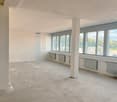 Large open-plan office space with white walls, concrete floor, and large windows providing natural light. The space appears to be in the process of renovation or construction, with visible heating units along the walls.