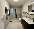 Bathroom: white tiled floor, white ceramic sink, modern shower stall, white toilet, mirror with lighting