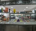 A restaurant kitchen with a counter, a glass display case with various food items, and a refrigerator. There are bottles, plates, and other items on the counter. The ceiling has lights and a surveillance camera. There are tables and chairs in the dining a