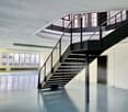 The image shows a modern, open office space with a large staircase in the center. The space has high ceilings, large windows, and a sleek, minimalist design. The staircase features a black metal frame and glass railings, creating a visually striking focal