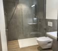 Modern bathroom with walk-in shower, toilet, sink, wooden flooring, and gray tiled walls.