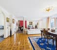 living room with wooden floor, white walls, white sofa, dining table, blue rug, chandelier, paintings, fireplace, windows with curtains